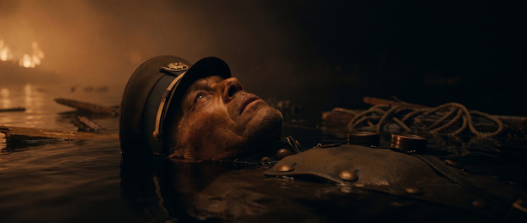 The Captain floating on his back in black night water, his face lit by distant fire through the fog.